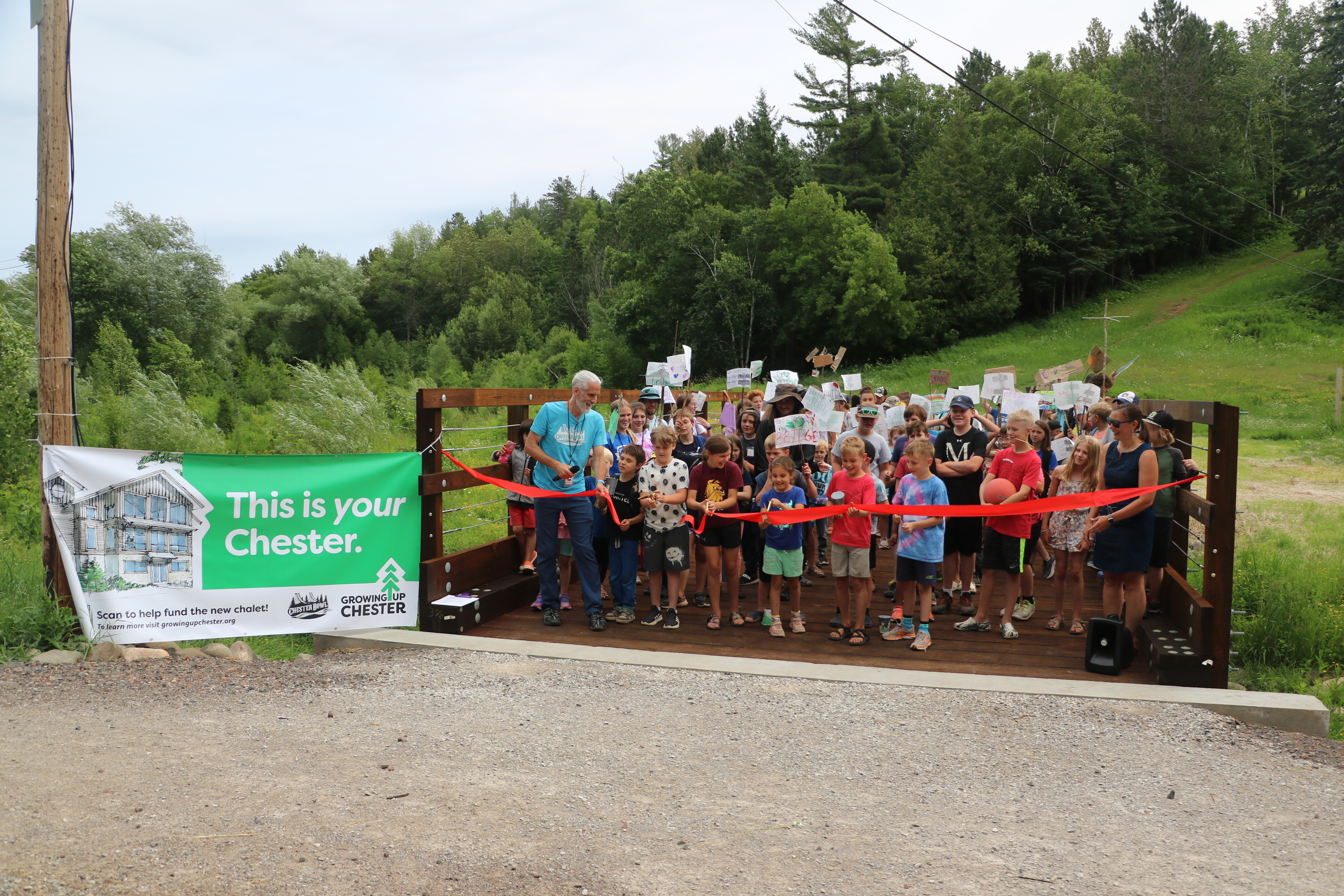 Photo from ribbon cutting event with a sign on left that says this is your Chester. There are one hundred kids on the bridge with five in front cutting a large red ribbon spanning the bridge. The background is lush and green and the kids are excited.